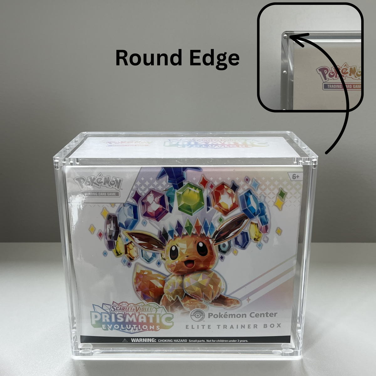 A clear acrylic Pokémon Center Elite Trainer Box displaying a Pokémon Prismatic Evolutions booster pack, with a graphic callout showing the rounded corner and a brand logo in the top left.