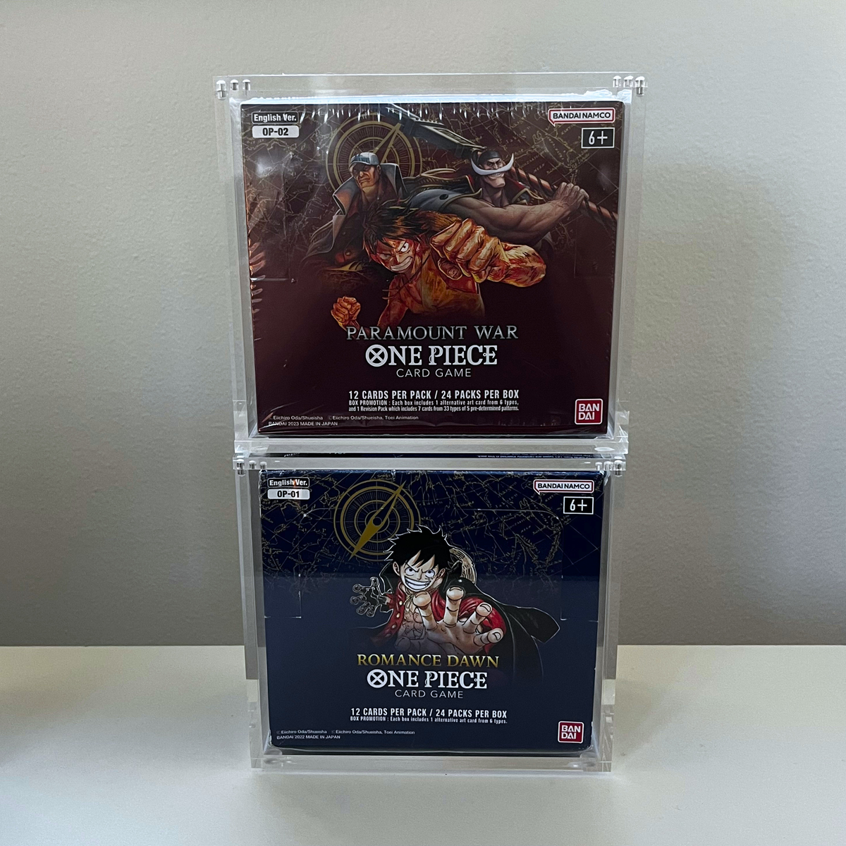 Two One Piece card game booster packs displayed in clear acrylic cases, stacked vertically on a white surface against a light gray wall. The top booster box is 'Paramount War' and the bottom booster box is 'Romance Dawn'.