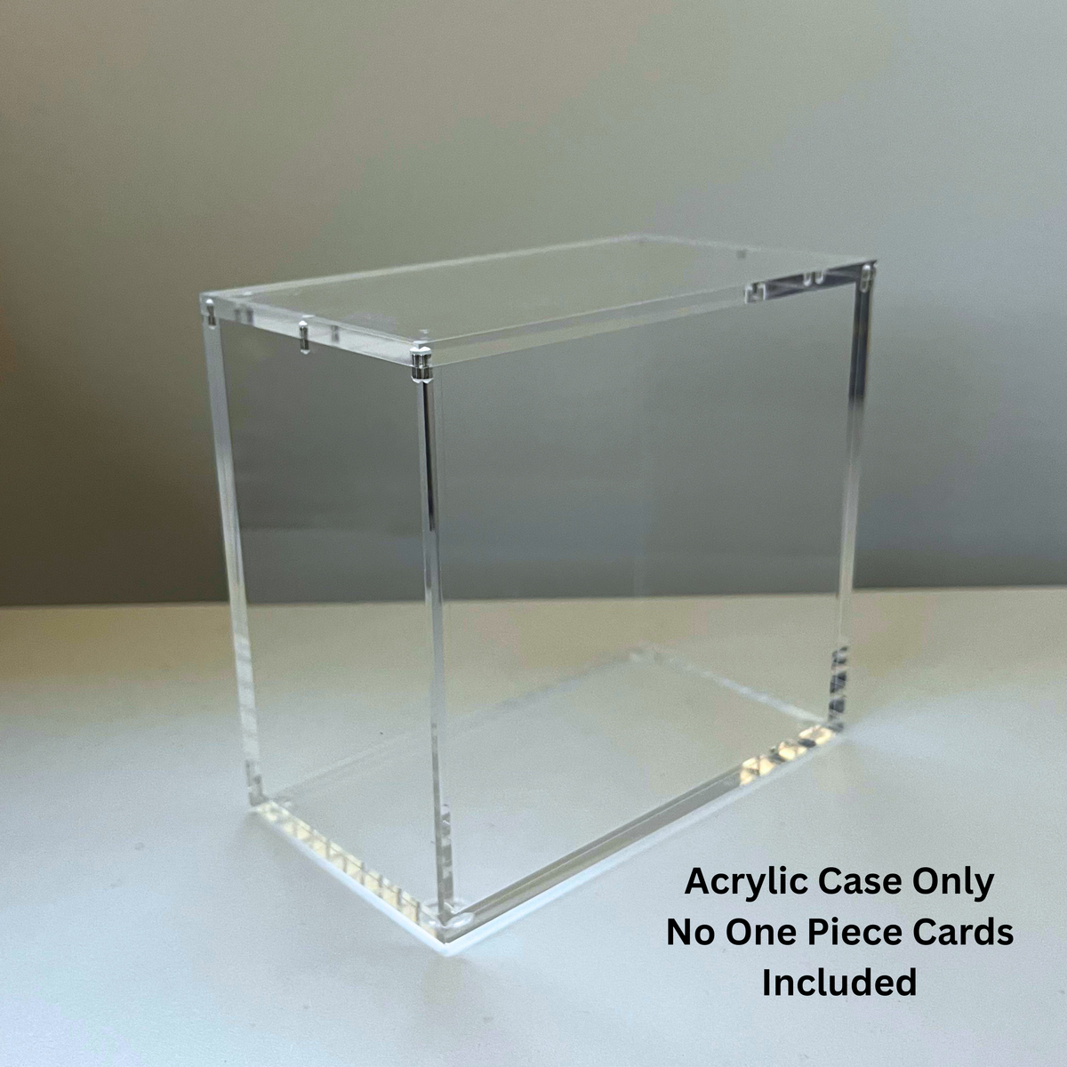 A clear acrylic display case for One Piece Booster Storage with a rectangular shape, shown on a light surface against a neutral gray background. 