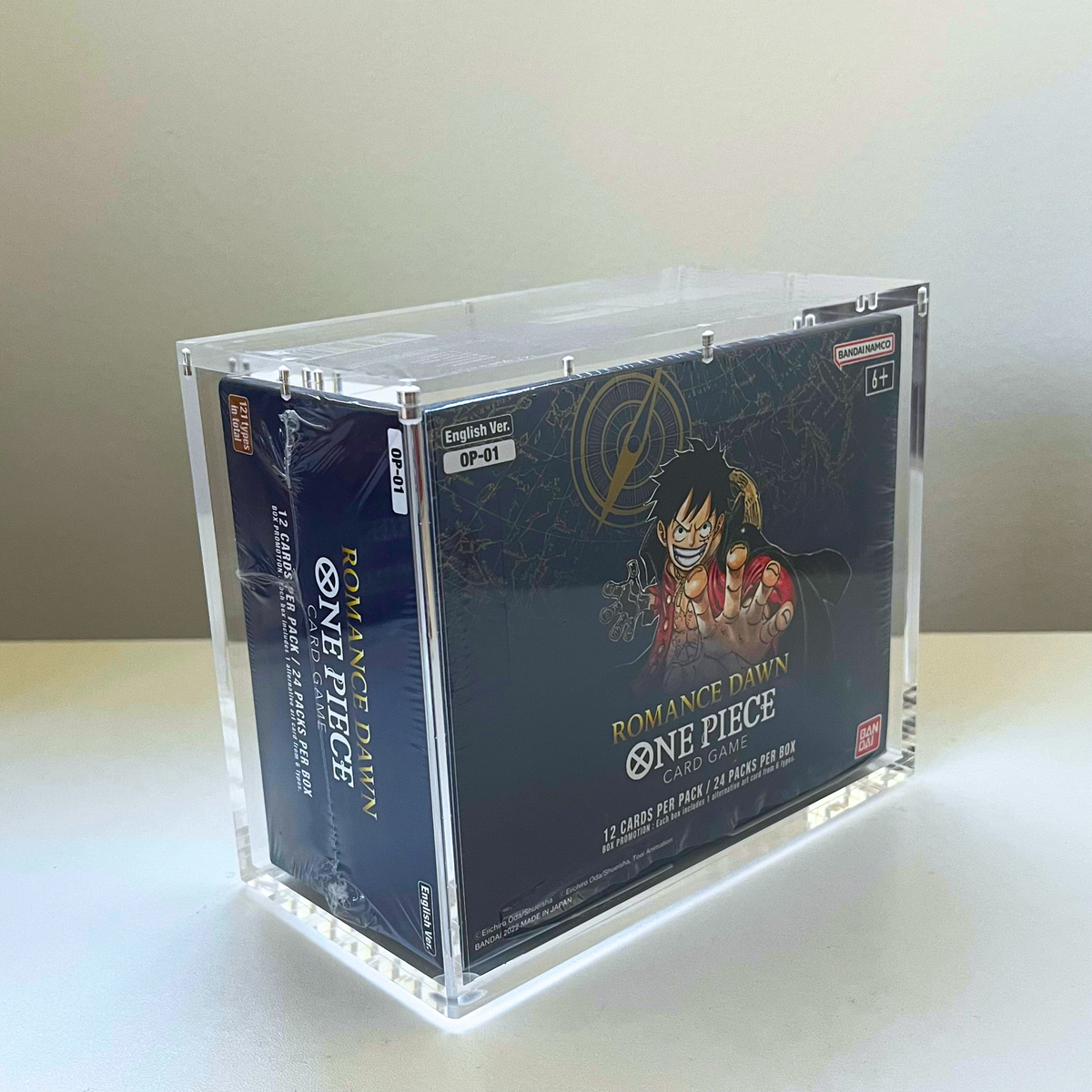 A clear acrylic display box holding a boxed One Piece card pack, photographed on a light tabletop against a plain wall. The visible product packaging shows One Piece branding and artwork, with a Gen Gaming watermark in the upper left.