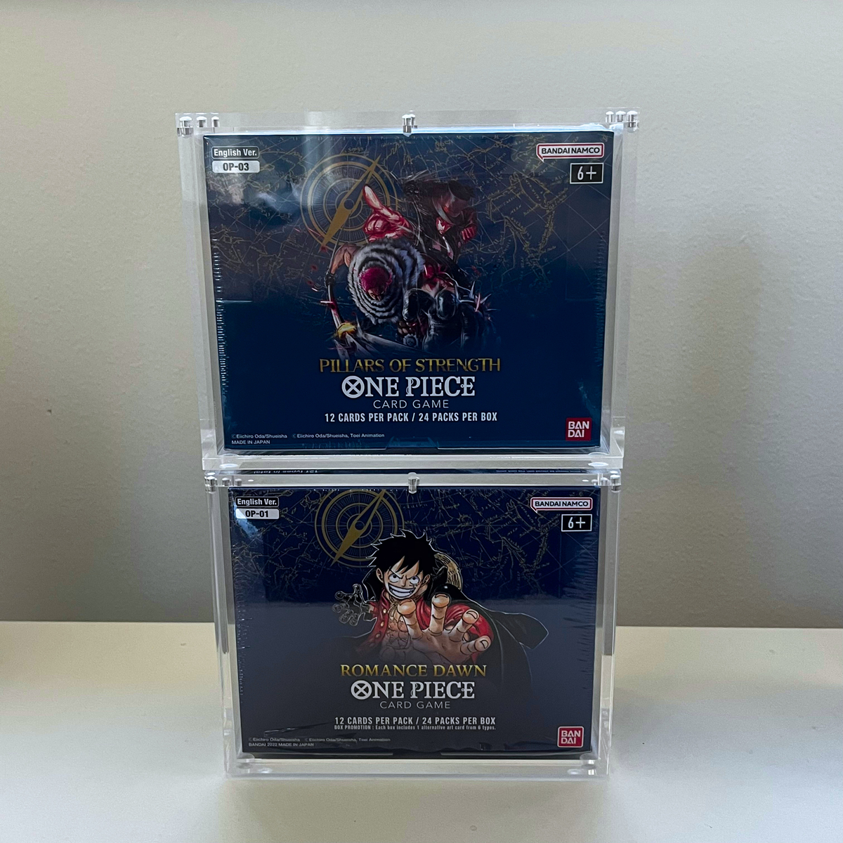 Two clear plastic display boxes stacked vertically on a white surface against a beige wall. The top box contains a One Piece OP-03 Pillars of Strength , and the bottom box contains a One Piece OP-01 Romance Dawn booster box.