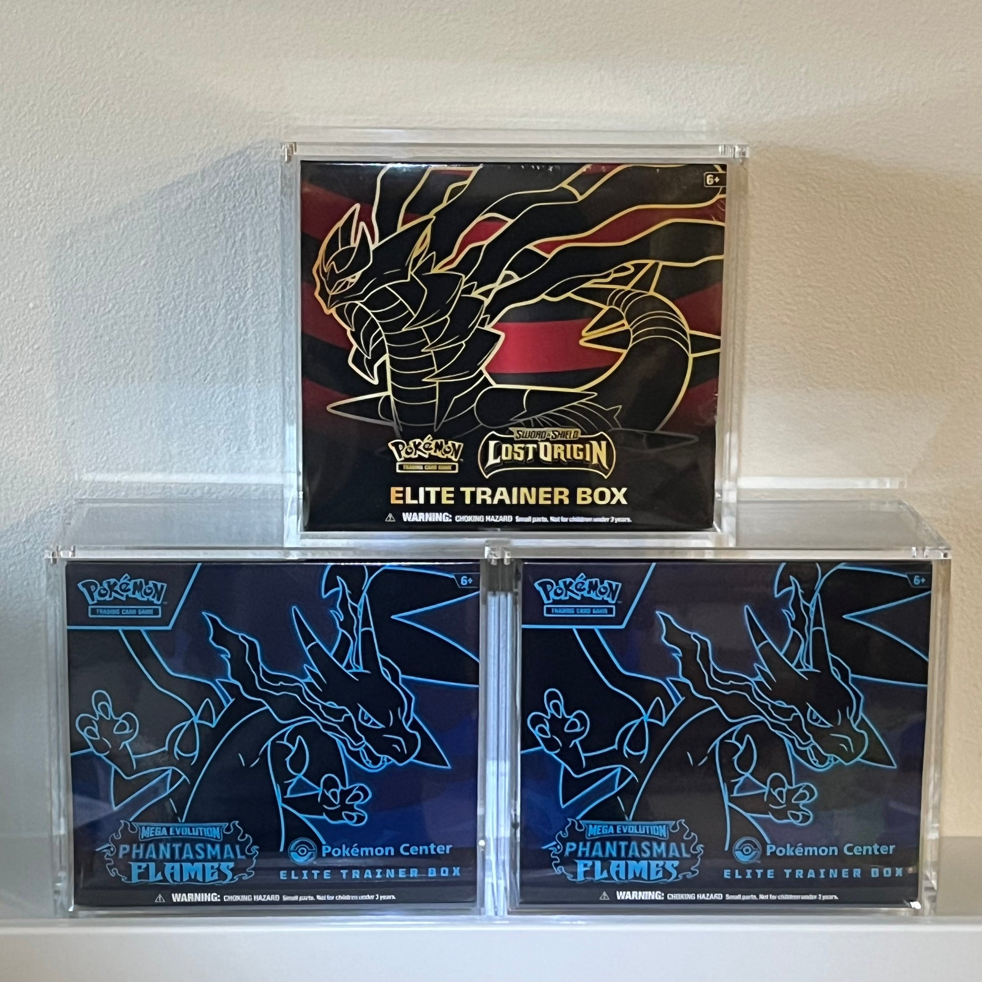 Pokémon Lost Origin and Pokemon Center Phantasmal Flames Elite Trainer Boxes stacked and on display in acrylic display cases.